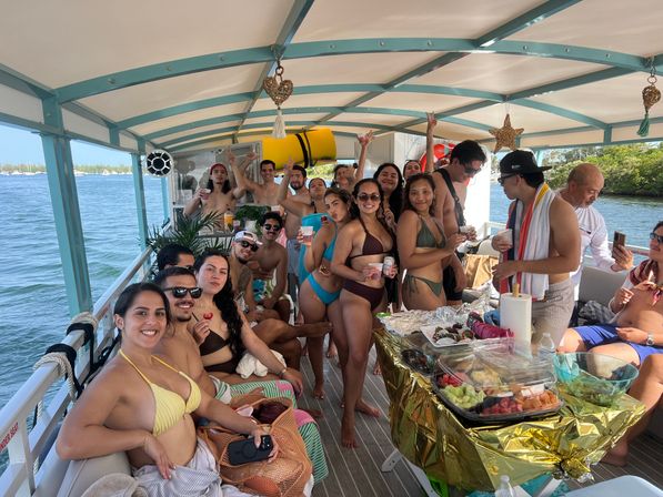 Group of friends in swimsuits partying on a covered pontoon boat in sunny coastal waters, smiling and holding drinks around a buffet table of fruit and snacks with teal canopy and shoreline in the background.
