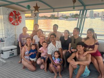 Smiling group of adults and children on a covered party boat at sunset, with a golden bay, moored yachts and a distant city skyline visible through the windows.