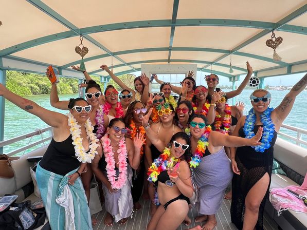 Group of women in colorful leis and novelty sunglasses partying and posing on a covered boat over turquoise tropical coastal waters.