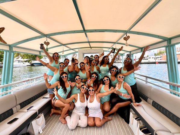Group of about 18 women in matching mint-green tank tops and white heart-shaped sunglasses cheering and posing on a covered teal-and-cream pontoon boat at a sunny marina with yachts and water in the background.