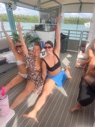 Three women in swimsuits smiling and doing splits with arms raised on a covered pontoon boat deck, turquoise coastal water and a tree-lined shoreline in the background — fun boat party scene.