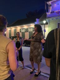 Nighttime historic-district street scene: a person in a leopard-print dress and heels talking to a small group under a lit balcony with string lights, shuttered windows, a lamp post and parked cars — lively nightlife atmosphere.