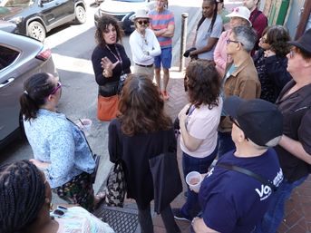Guide animatedly gesturing to a group of adults on a sunny downtown walking tour along a brick sidewalk beside parked cars