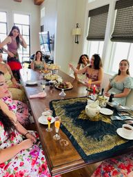 Sunlit home tea party: a group of women gathered around a long wooden table in a modern dining room, enjoying tea and champagne flutes with tiered sandwiches, pastries and chocolate-covered strawberries.