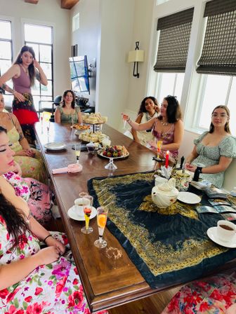 Sunlit home tea party: a group of women gathered around a long wooden table in a modern dining room, enjoying tea and champagne flutes with tiered sandwiches, pastries and chocolate-covered strawberries.