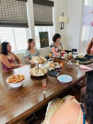 Sunlit indoor afternoon tea in a bright dining room: four women in floral dresses around a wooden table with tiered finger sandwiches, pastries, chocolate-covered strawberries, a teapot and champagne flutes.