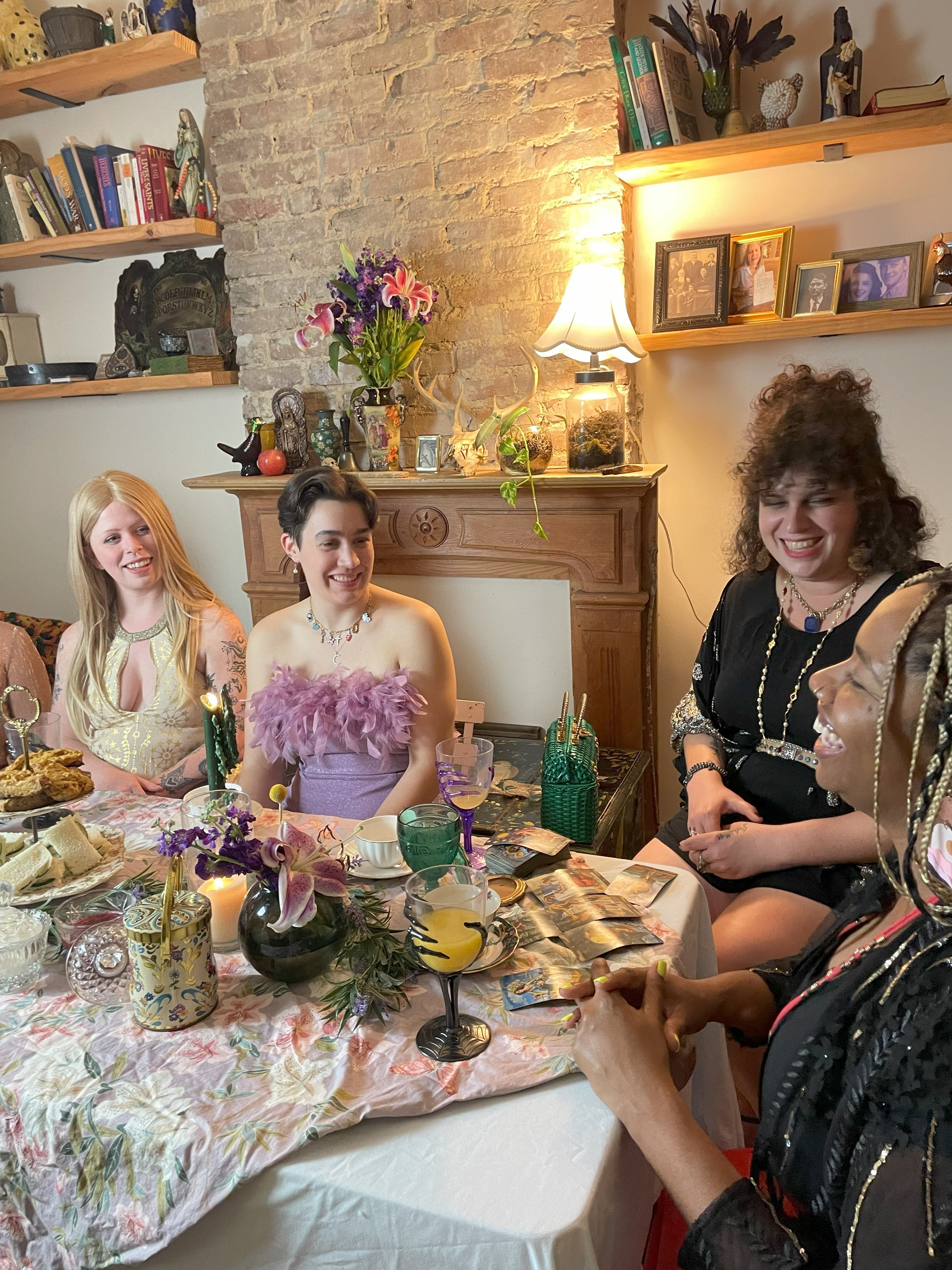 Four friends laughing around a floral-covered table in a cozy vintage living room, enjoying afternoon tea and pastries with colorful glassware, a lily bouquet on an exposed-brick mantel and tarot cards.