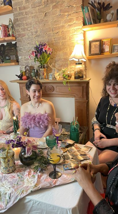 Four friends laughing around a floral-covered table in a cozy vintage living room, enjoying afternoon tea and pastries with colorful glassware, a lily bouquet on an exposed-brick mantel and tarot cards.