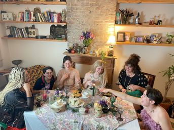 Six friends laughing around a cozy apartment dining table set with a floral tablecloth, tea set, candles and snacks, with an exposed brick fireplace, bookshelves and framed photos in the background.