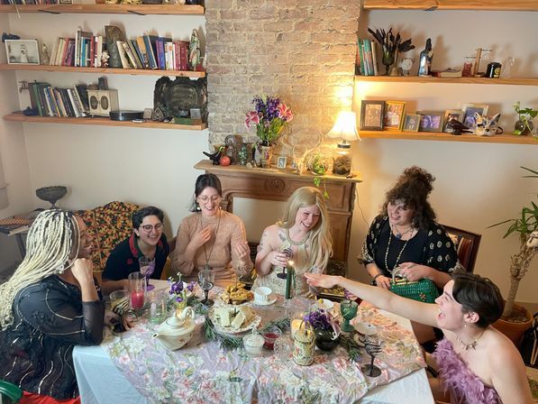 Six friends laughing around a cozy apartment dining table set with a floral tablecloth, tea set, candles and snacks, with an exposed brick fireplace, bookshelves and framed photos in the background.