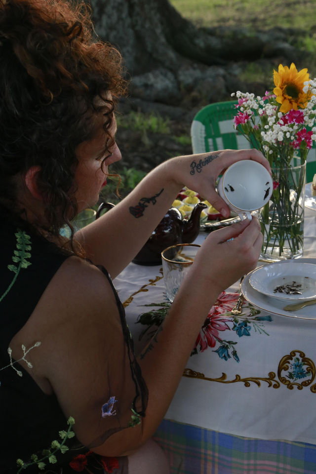Curly-haired woman at an outdoor garden tea party holding a vintage teacup upside down to inspect tea leaves, with a floral tablecloth, sunflower bouquet and teapot nearby.