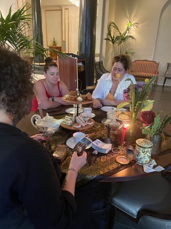 Cozy afternoon tea in an elegant hotel lobby: three people gathered around a round table with a tiered tray of sandwiches and pastries, vintage teapot and cups, lit candle and floral centerpiece while one person handles tarot cards for a playful tea-time reading.