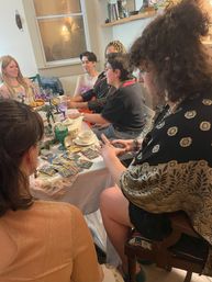 Cozy indoor tarot tea party with friends gathered around a dining table, tarot cards spread among teapot, cups, pastries and colorful glassware
