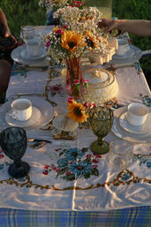 Outdoor garden tea-party table at golden hour with vintage white teacups and plates, colored goblets, a vase of sunflowers and baby's breath, a tiered stand of finger sandwiches and a floral tablecloth.