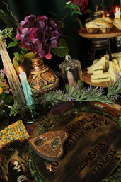 Victorian-style spooky tea setting with a wooden Ouija board and ornate planchette, lit blue candle, purple hydrangea in a patterned vase, lavender sprigs, glass apothecary jar, and a tiered tray of scones and cucumber tea sandwiches.