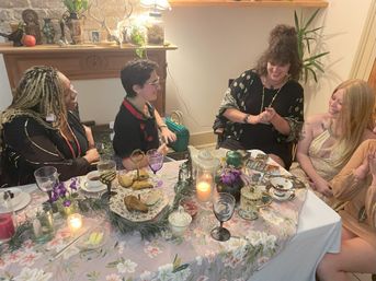 Cozy indoor tea party in a living room: five people laughing around a floral-covered table set with a teapot, teacups, pastries, candles, flowers and tarot cards during a friendly reading.