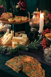 Candlelit vintage tea spread with a two-tier wooden stand of crustless finger sandwiches and mini tarts, brass teapot, red grapes, a burning pillar candle, sprigs of greenery and colorful tarot-style cards on dark velvet.
