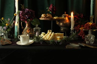 Elegant vintage afternoon tea still life: teacup and saucer, finger sandwiches and scones on a tiered stand, lit taper candles, grapes and floral arrangements on a dark velvet table.
