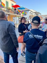 Group chatting on a sunny historic downtown street with balcony cafes, rainbow pride flags, and a visible volunteer shirt