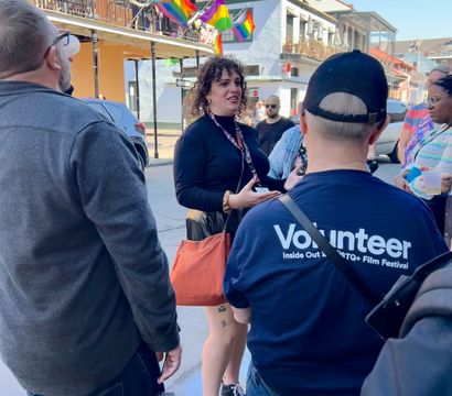 Group chatting on a sunny historic downtown street with balcony cafes, rainbow pride flags, and a visible volunteer shirt