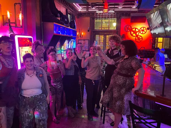 Friends cheering with drinks in a neon-lit bar arcade, standing by colorful gaming machines and a long wooden bar with decorative gear wall art.