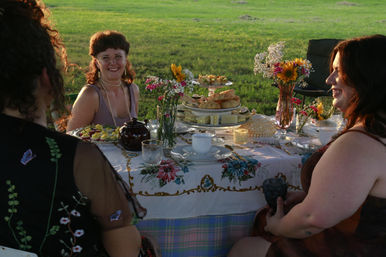 Three women enjoying an outdoor tea party in a sunlit meadow, floral tablecloth set with teapot and teacups, tiered stand of scones and sandwiches, and vases of sunflowers and wildflowers.