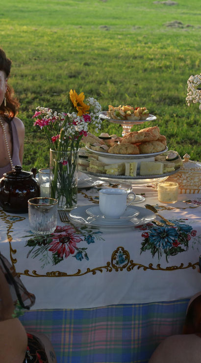 Three women enjoying an outdoor tea party in a sunlit meadow, floral tablecloth set with teapot and teacups, tiered stand of scones and sandwiches, and vases of sunflowers and wildflowers.