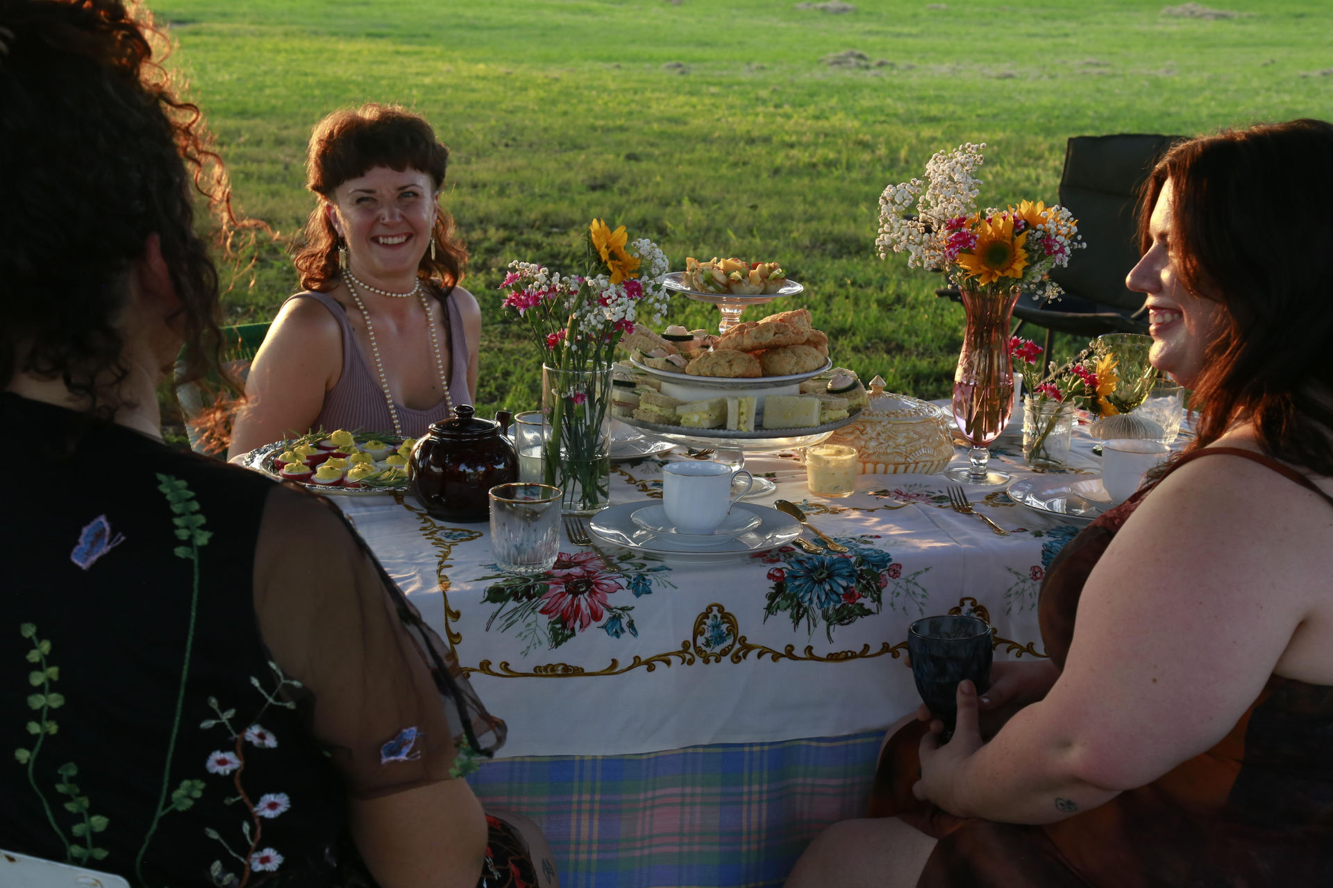 Three women enjoying an outdoor tea party in a sunlit meadow, floral tablecloth set with teapot and teacups, tiered stand of scones and sandwiches, and vases of sunflowers and wildflowers.
