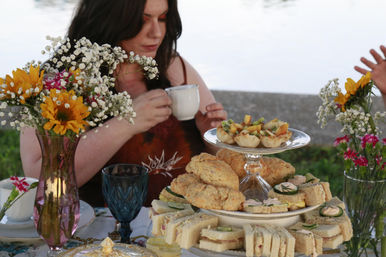 Lakeside afternoon tea scene with a woman holding a teacup, a tiered tray of finger sandwiches, scones and mini tarts, blue goblet, and vases of sunflowers and baby's breath