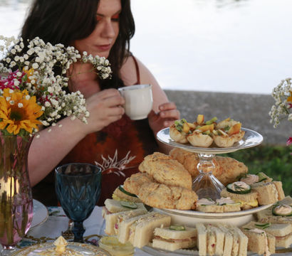 Lakeside afternoon tea scene with a woman holding a teacup, a tiered tray of finger sandwiches, scones and mini tarts, blue goblet, and vases of sunflowers and baby's breath