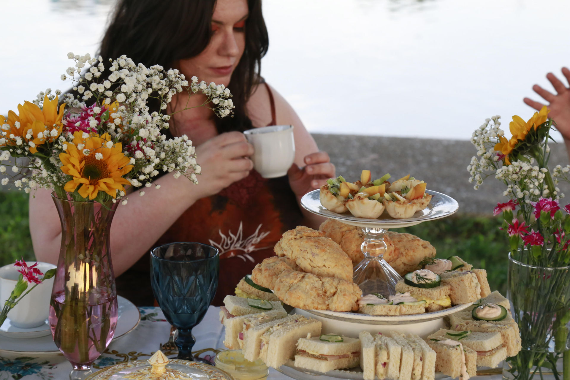 Lakeside afternoon tea scene with a woman holding a teacup, a tiered tray of finger sandwiches, scones and mini tarts, blue goblet, and vases of sunflowers and baby's breath
