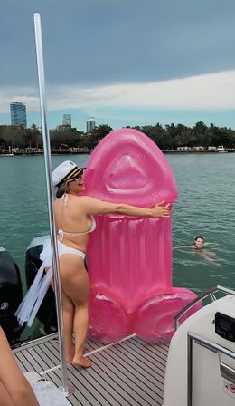 Woman in white bikini and captain's hat hugging a large pink novelty inflatable on a boat in a turquoise bay with palm-lined shore and city skyline