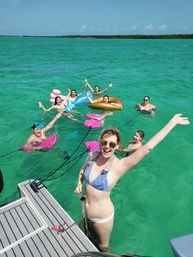 Group of friends in swimsuits splashing and smiling in clear turquoise water off a boat, floating on colorful inflatables and waving on a sunny tropical day