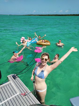 Group of friends in swimsuits splashing and smiling in clear turquoise water off a boat, floating on colorful inflatables and waving on a sunny tropical day