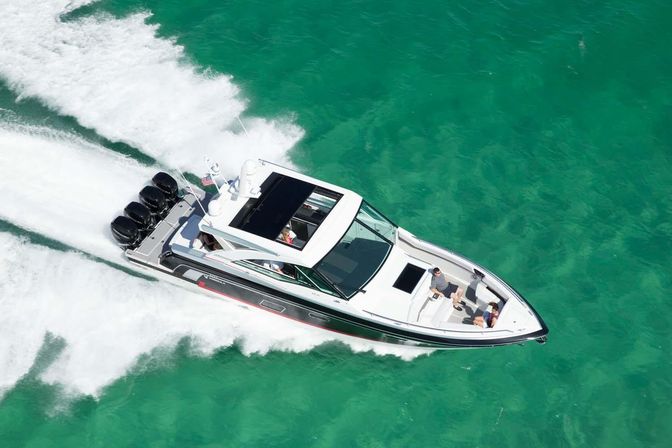 Aerial view of a white luxury speedboat with four outboard engines zipping across clear turquoise water, leaving a white wake while passengers relax on the bow.