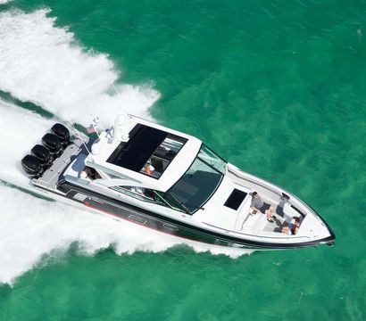 Aerial view of a white luxury speedboat with four outboard engines zipping across clear turquoise water, leaving a white wake while passengers relax on the bow.