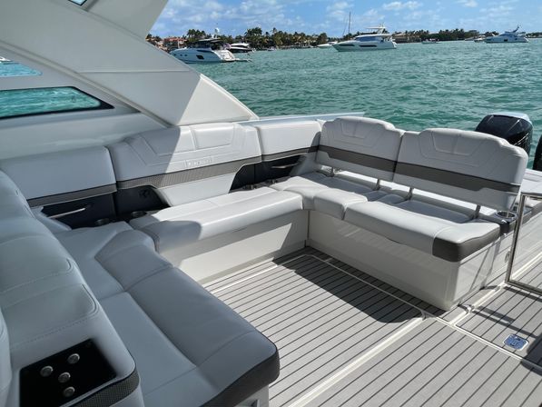 Luxury white L-shaped boat seating and gray teak-style deck on a sunny day, turquoise water with anchored yachts and a palm-lined shoreline in the background.