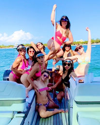 Group of women in colorful swimsuits partying on a motorboat, one posing on a dance pole, turquoise ocean and palm-lined tropical coastline under a clear blue sky