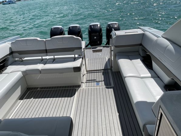 Sunlit luxury boat aft deck with gray cushioned seating, synthetic teak-style flooring, gated swim platform and four black outboard engines backed by turquoise open water.