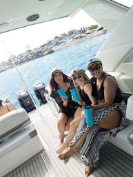 Three friends in captain hats smiling on a sunny yacht deck, holding bright blue tumblers with turquoise water and a marina in the background.