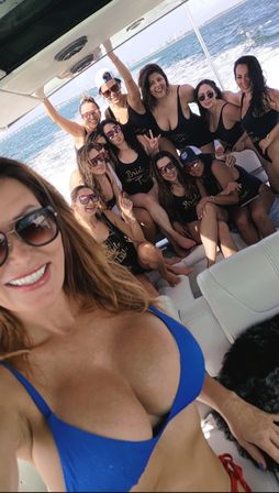 Selfie of a smiling woman in a blue bikini with sunglasses and a group of friends in matching 'bride tribe' swimsuits enjoying a sunny boat cruise on coastal waters with the ocean and skyline in the distance.