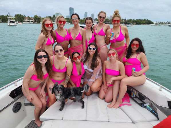 Dozen women in matching hot-pink bikinis and sunglasses posing for a boat party on a turquoise bay with a coastal skyline and a small black-and-white dog.