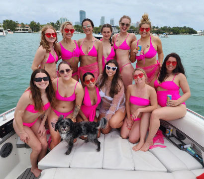 Dozen women in matching hot-pink bikinis and sunglasses posing for a boat party on a turquoise bay with a coastal skyline and a small black-and-white dog.