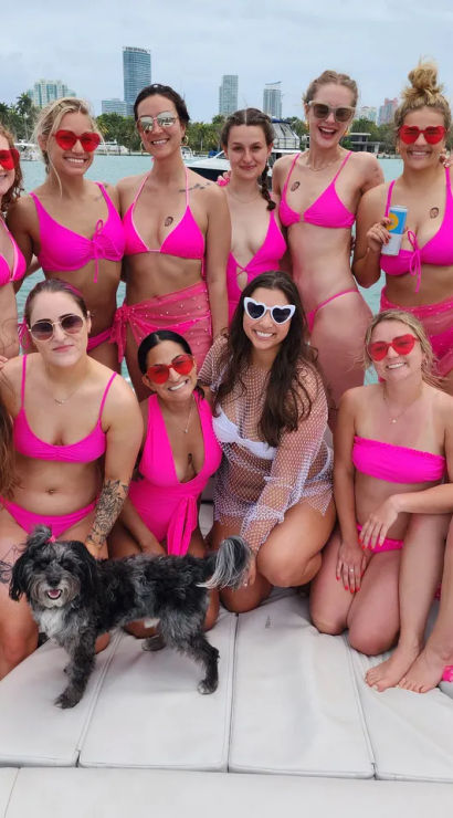 Dozen women in matching hot-pink bikinis and sunglasses posing for a boat party on a turquoise bay with a coastal skyline and a small black-and-white dog.
