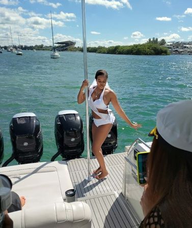 Woman in a white swimsuit posing on a motorboat deck by a dance pole, turquoise coastal water with sailboats and mangrove shoreline under a sunny blue sky.
