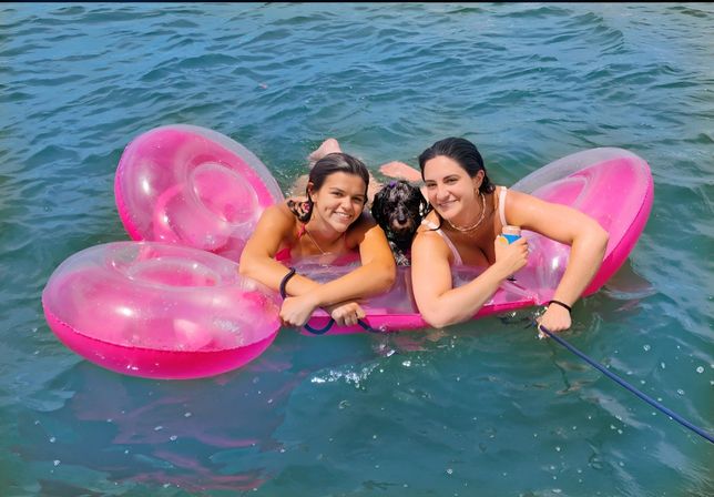 Two friends smiling on a bright pink inflatable float with a small black dog, holding a drink and relaxing on a sunny lake.