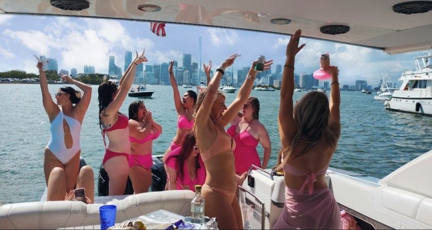 Bachelorette boat party: group of women in pink swimsuits and bikinis dancing and raising drinks on a yacht with a sunny city skyline and boats in the harbor
