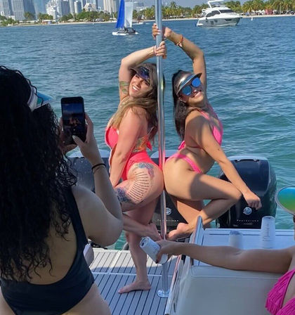 Two women in bright pink bikinis playfully posing on a pole aboard a yacht while a friend snaps a photo, turquoise water with palm-lined shore and coastal city skyline in the background.