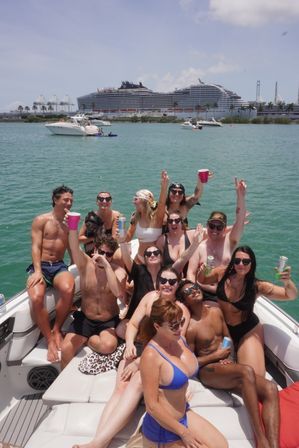 Group of friends partying on a sunlit motorboat, holding drinks in turquoise harbor waters with a large cruise ship and palm-lined port in the background