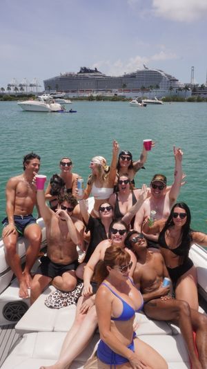 Group of friends partying on a sunlit motorboat, holding drinks in turquoise harbor waters with a large cruise ship and palm-lined port in the background
